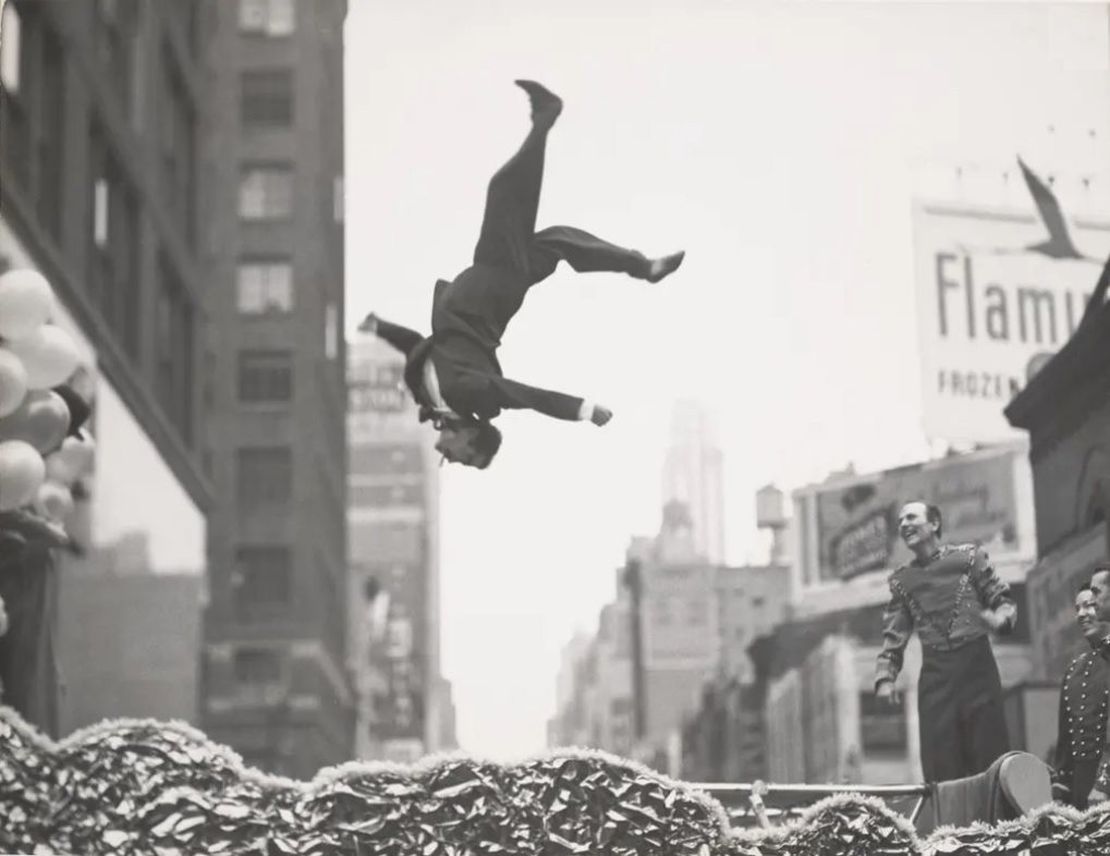 Photo Noir & Blanc de Garry Winogrand représentant un homme en costume faisant une pirouette en l'air devant l'air ébahi de quelques spectateurs (initiationphoto.com)