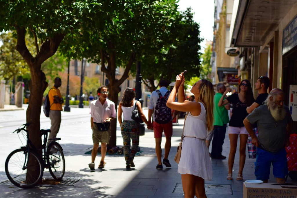 Photo couleur anonyme représentant une femme arrêtée sur un trottoir en ville et faisant la photo du ciel à travers les arbres (initiationphoto.com)