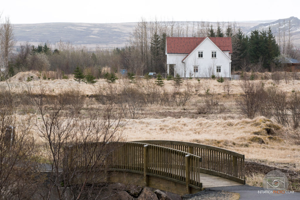 Photo de paysage en couleur, prise au téléobjectif représentant une maison et un petit pont de bois dans les plaines islandaises (initiationphoto)