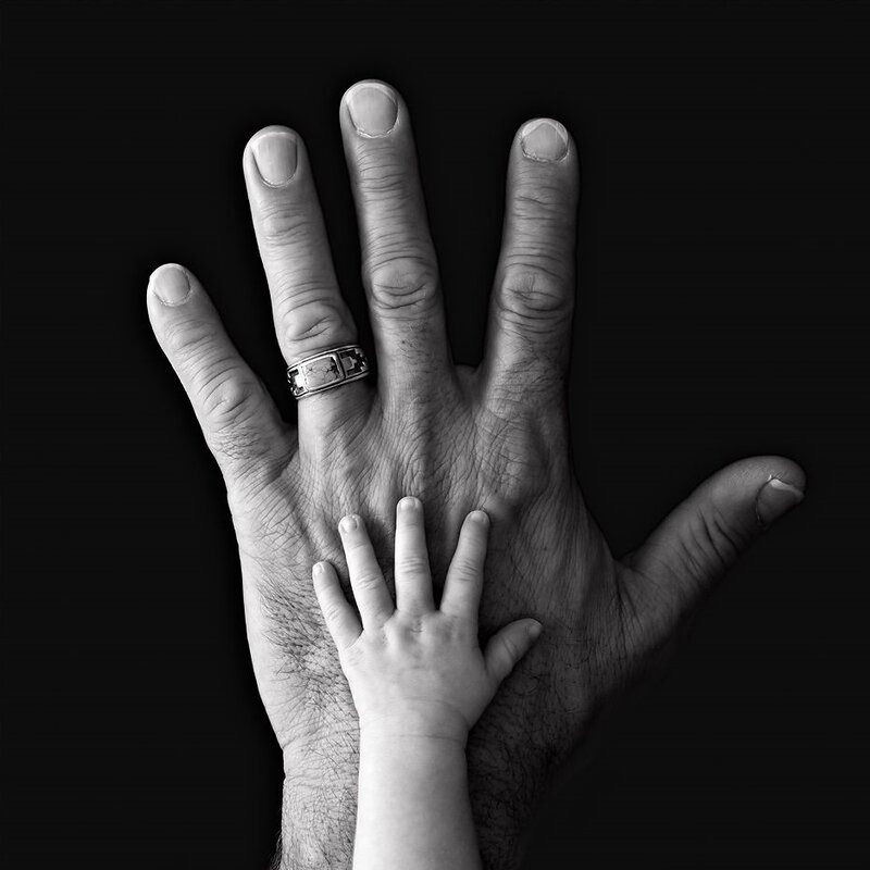Photo en noir et blanc de deux mains, celle d'un homme sur laquelle est posée celle d'un petit enfant, créant un contraste de taille.