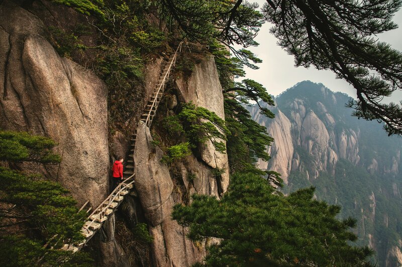 Photo couleur d'un paysage de montagne montrant une escalier sur une paroie rocheuse, sur lequel se trouve un homme avec un pull rouge